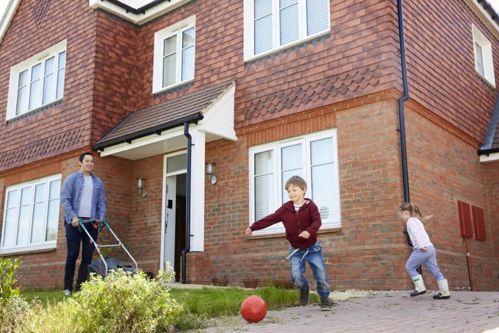 A father mowing the lawn while two children play football outside a red-brick family home